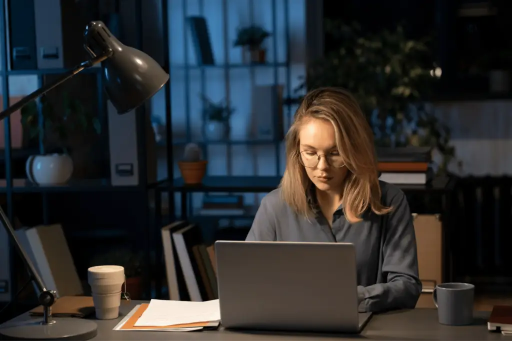 a professional women sitting and facing her laptop with spotlight on her face and a cup of coffee within her desk in a library set up
