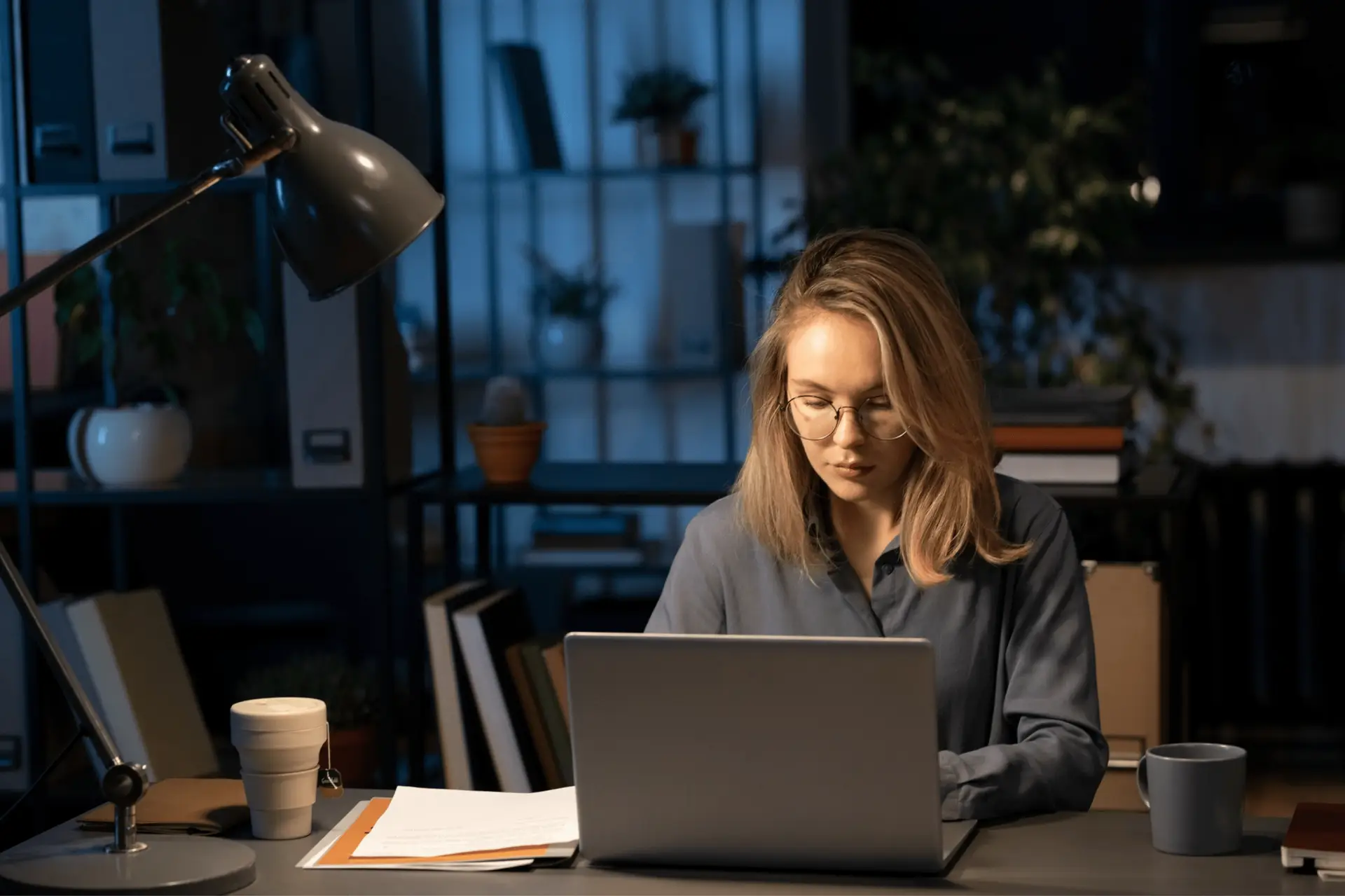 a professional women sitting and facing her laptop with spotlight on her face and a cup of coffee within her desk in a library set up