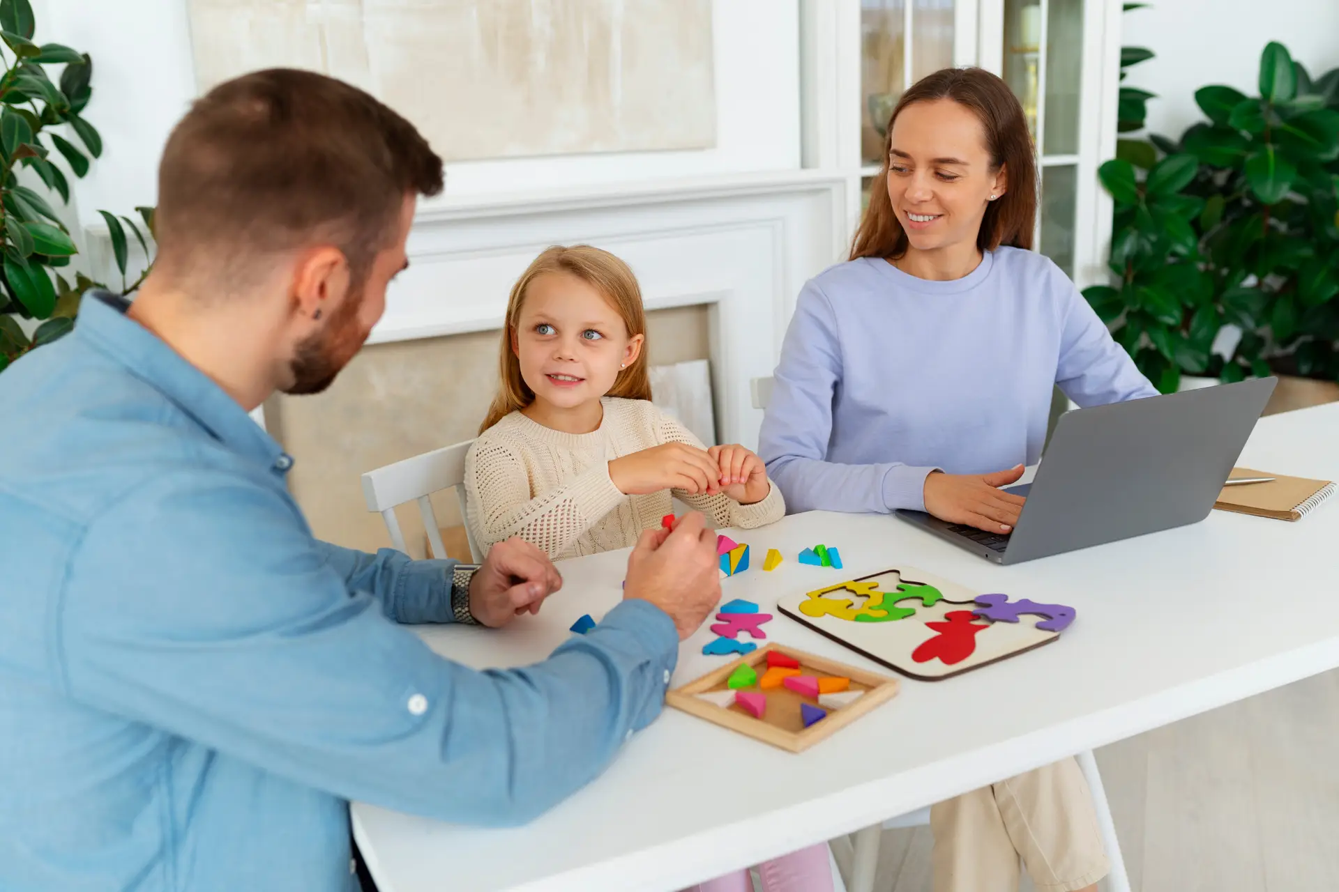 a discussion of a family - father facing his daughter in a table while playing her craft and arts with her mom behind working in a laptop and facing to her daughter