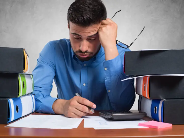 a stressed guy facing his paperworks handling his pen and eyeglass with a lot of books behind for review